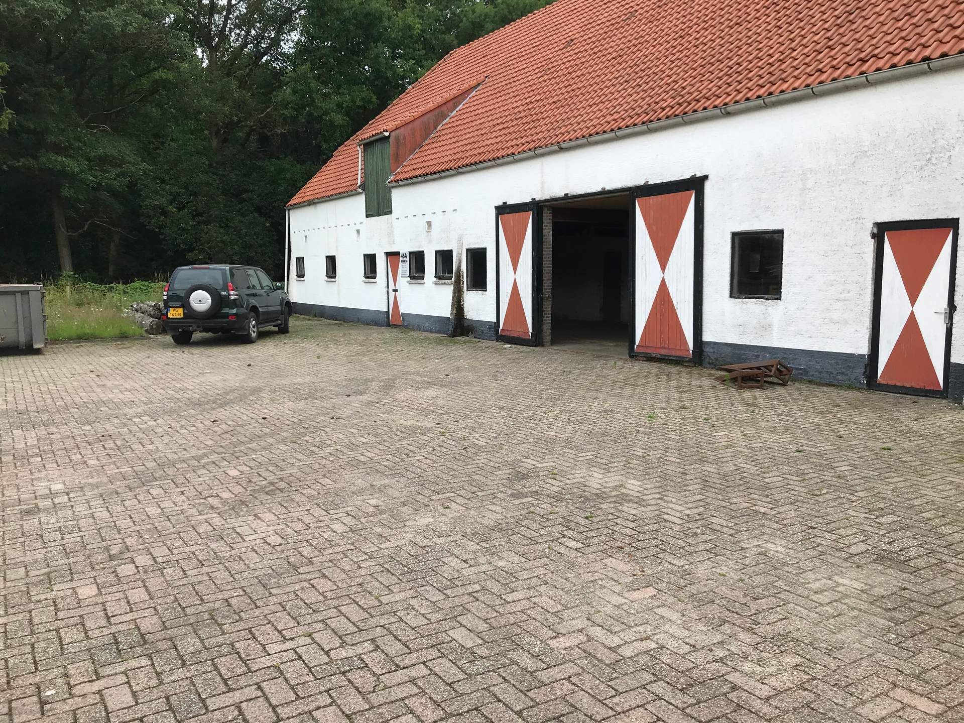 Witte boerderij met rood dak en schuurdeuren naast een geparkeerde zwarte auto op een geplaveide binnenplaats.