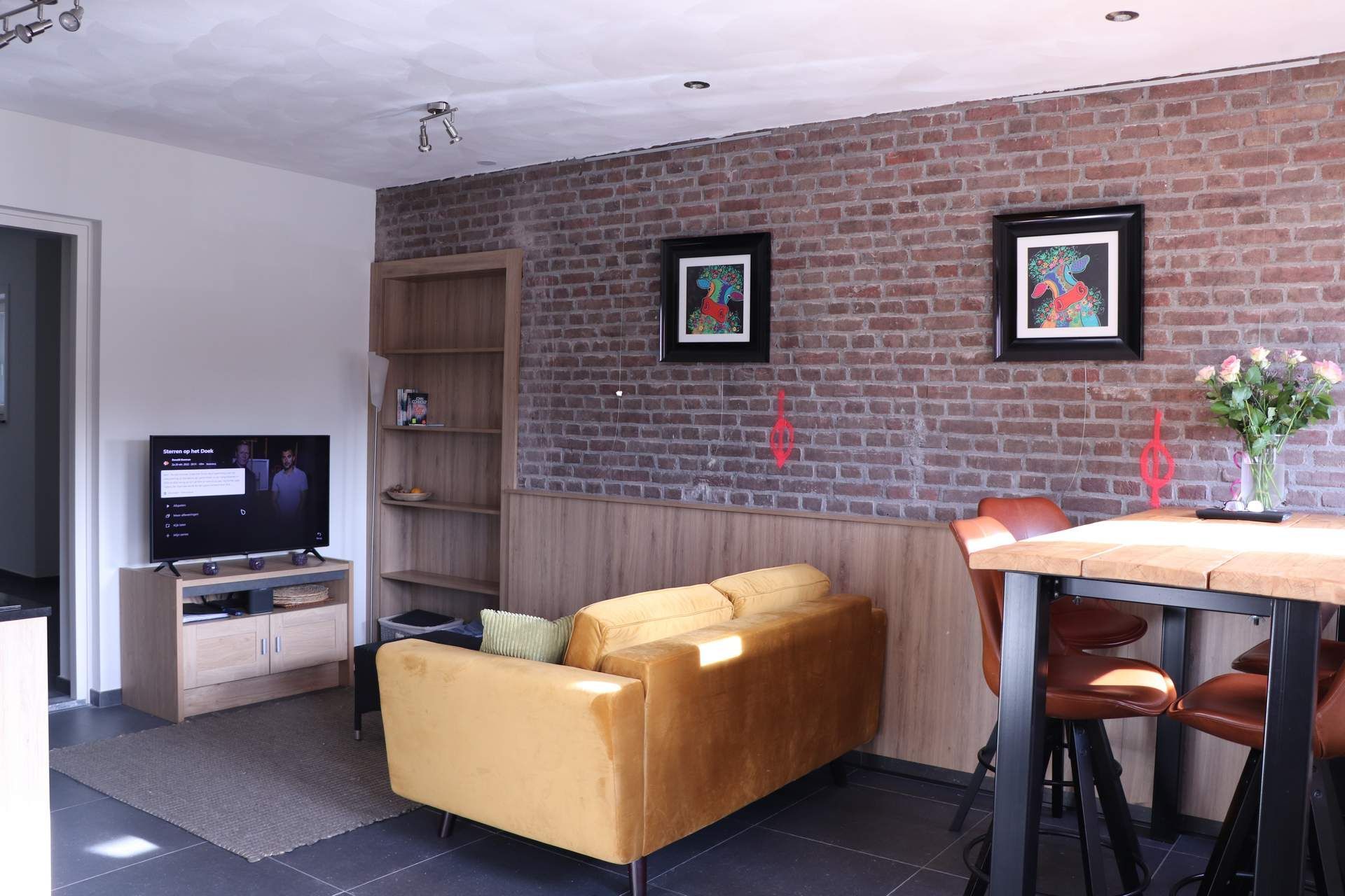 Modern living room with brick wall, yellow sofa, TV stand, framed art, and bar stools by a dining table