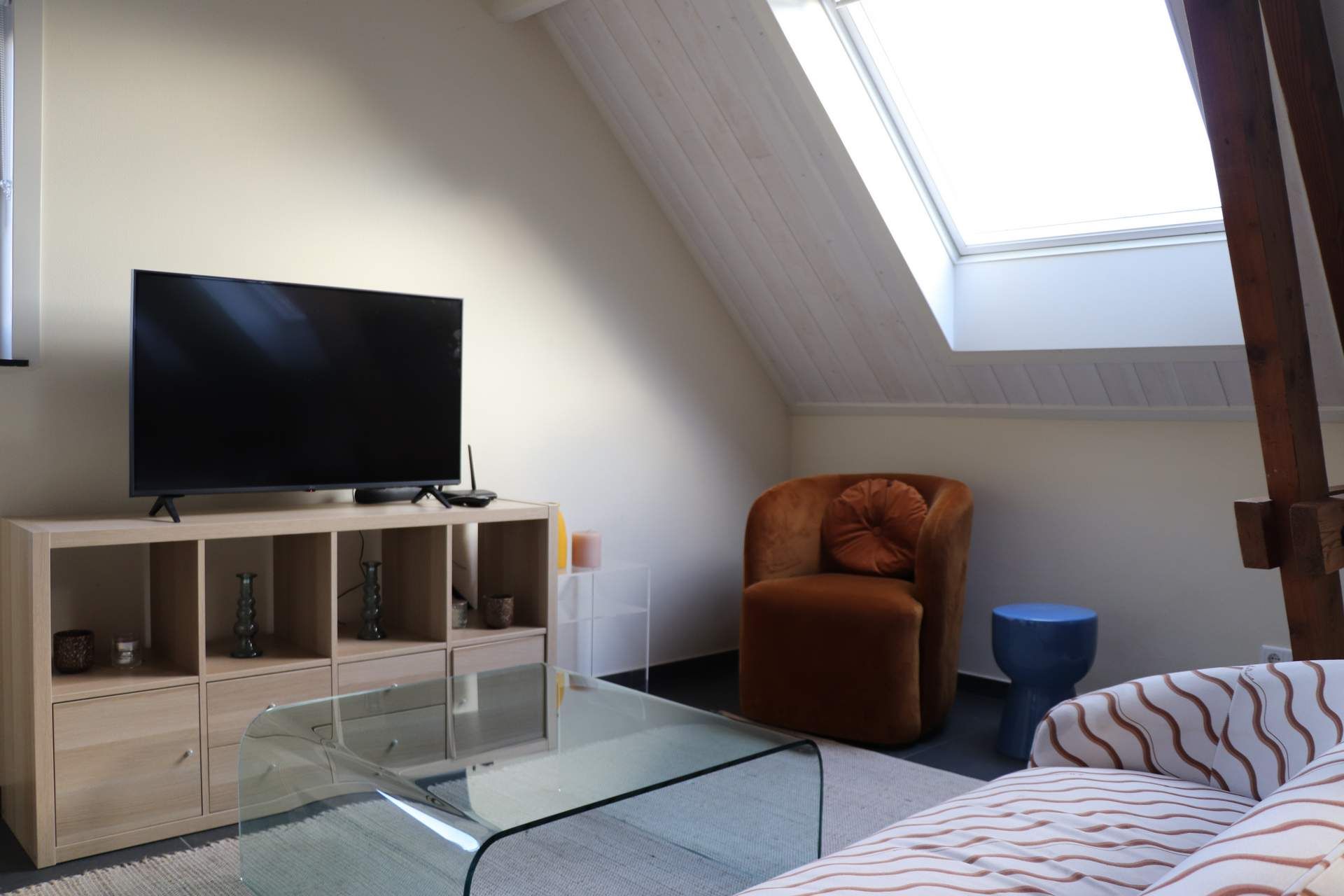 Cozy attic bedroom with TV on cabinet, brown chair, skylight, glass table, and striped bedspread