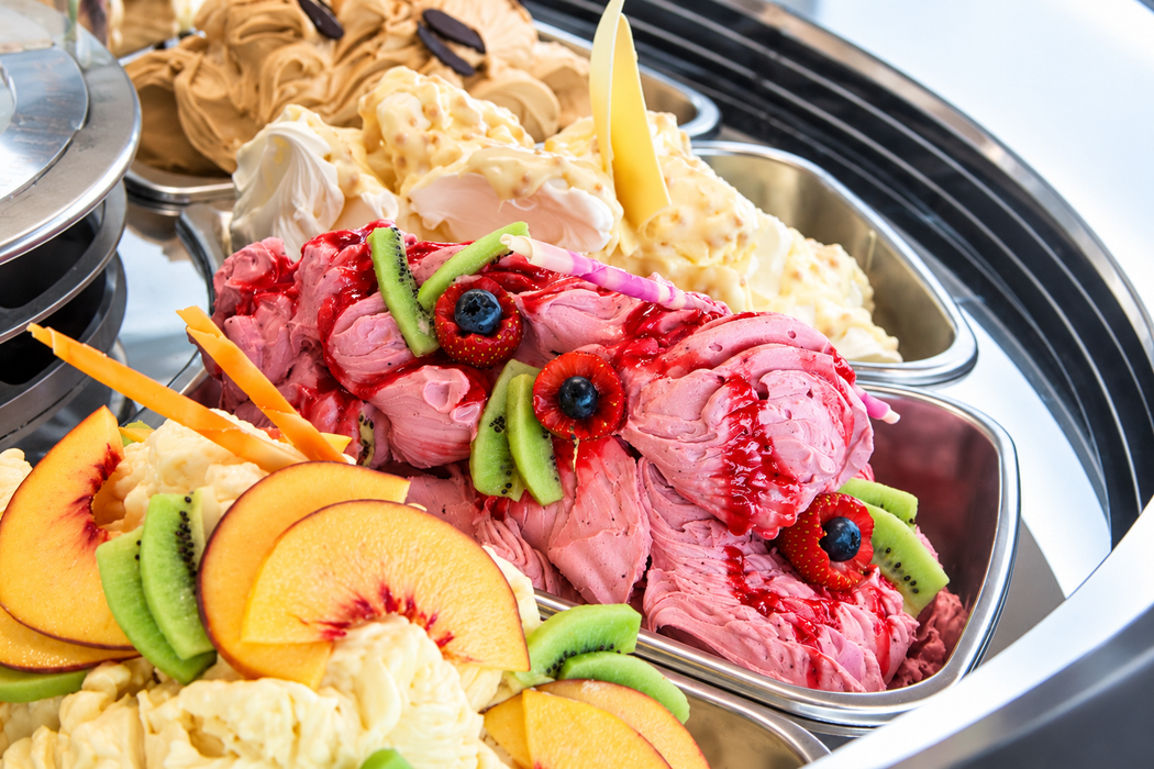 Gelato colorato guarnito con frutta in una vaschetta di metallo, con kiwi, pesca, frutti di bosco e panna montata, esposto in vetrina