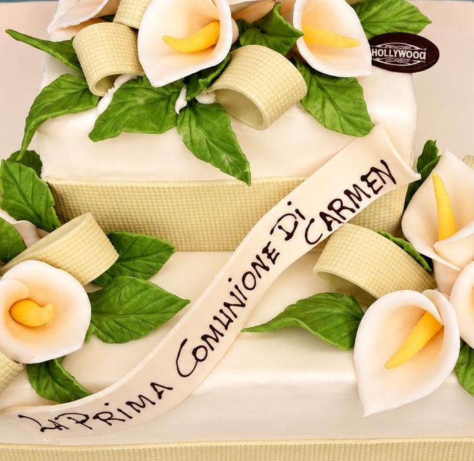 Torta bianca decorata con fiori di panna, foglie verdi e un nastro con la scritta “Prima Comunione di Carmen”