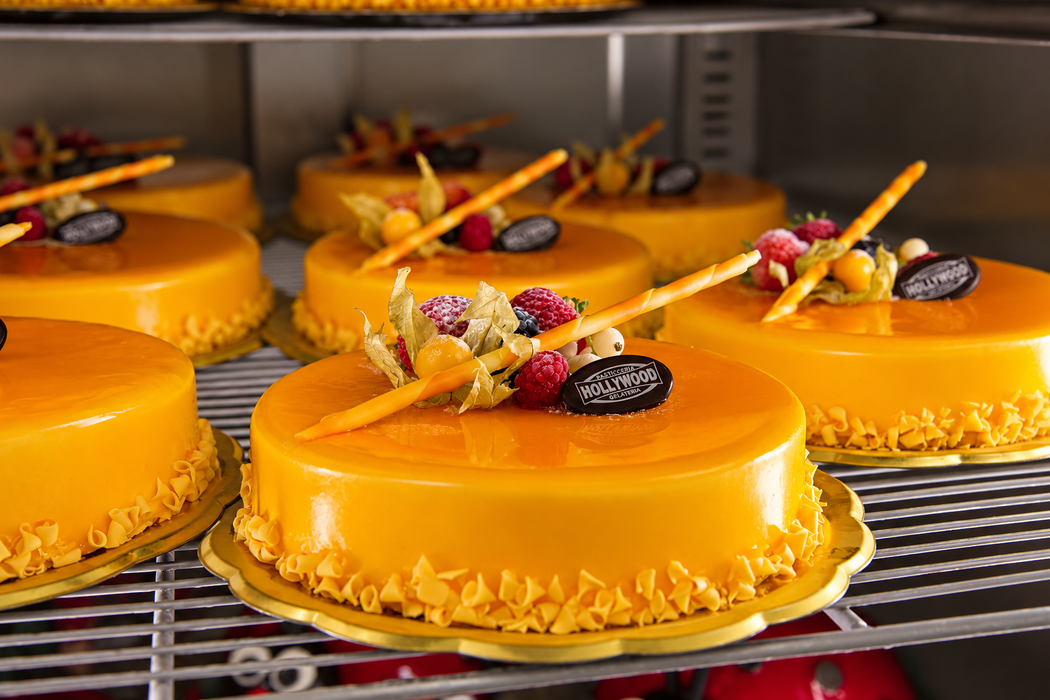 Dolci glassati all’arancia, decorati con frutti di bosco e cioccolato, su un ripiano metallico in una vetrina frigorifera