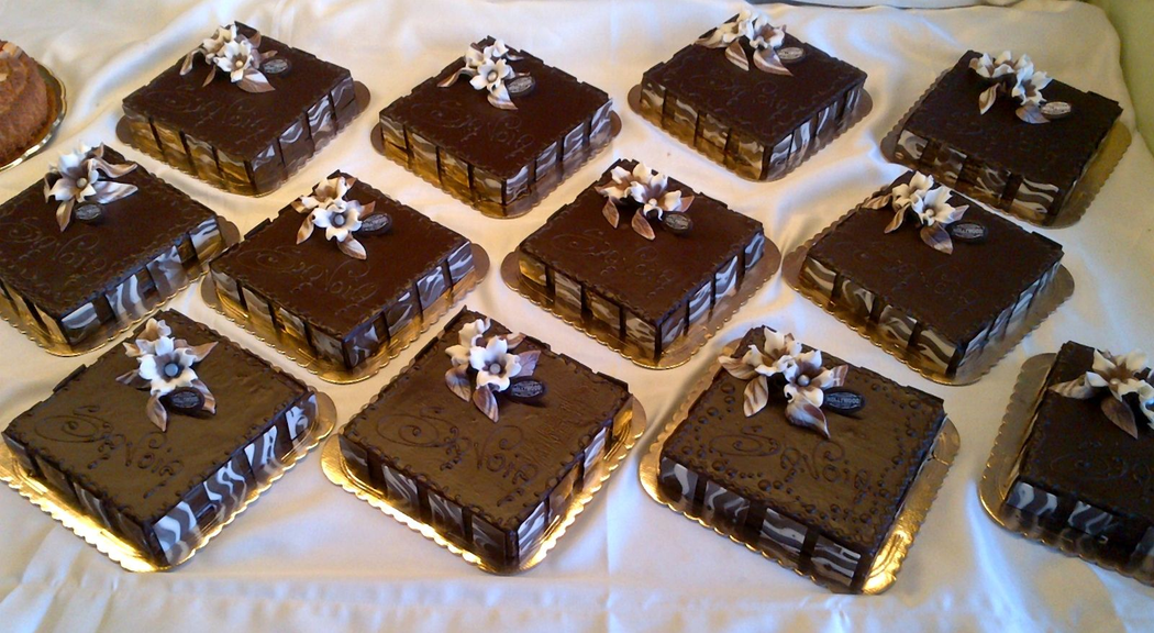 Diverse torte quadrate ricoperte di cioccolato su vassoi dorati, ciascuna decorata con fiori bianchi e viola.