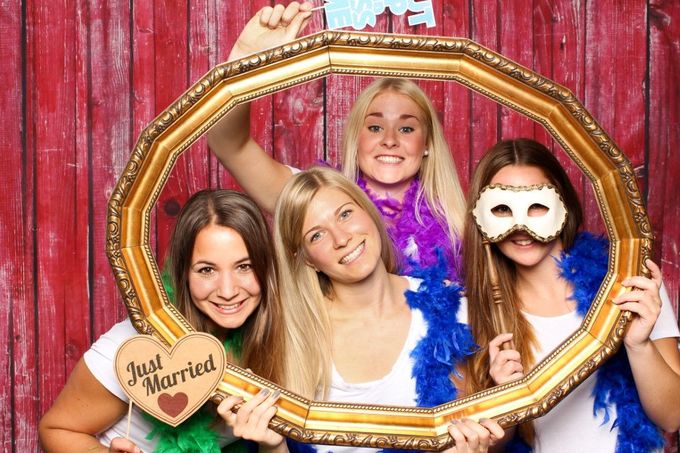 Four women posing in a photo booth, holding a gold frame and props, smiling.