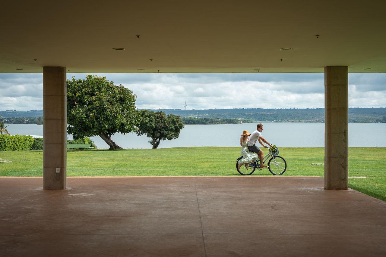 Homem anda de bicicleta com um passageiro ao longo de uma orla gramada. Vista emoldurada por um prédio bege. Céu e água azuis.