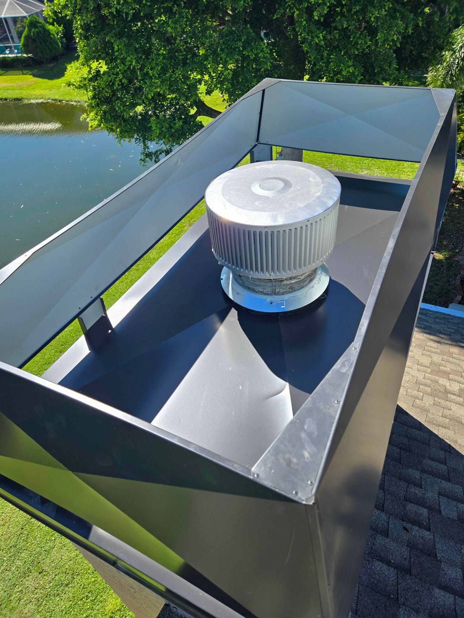 A silver chimney cap with a mesh screen sits centered on a black, sloped metal roof assembly against a pond background.