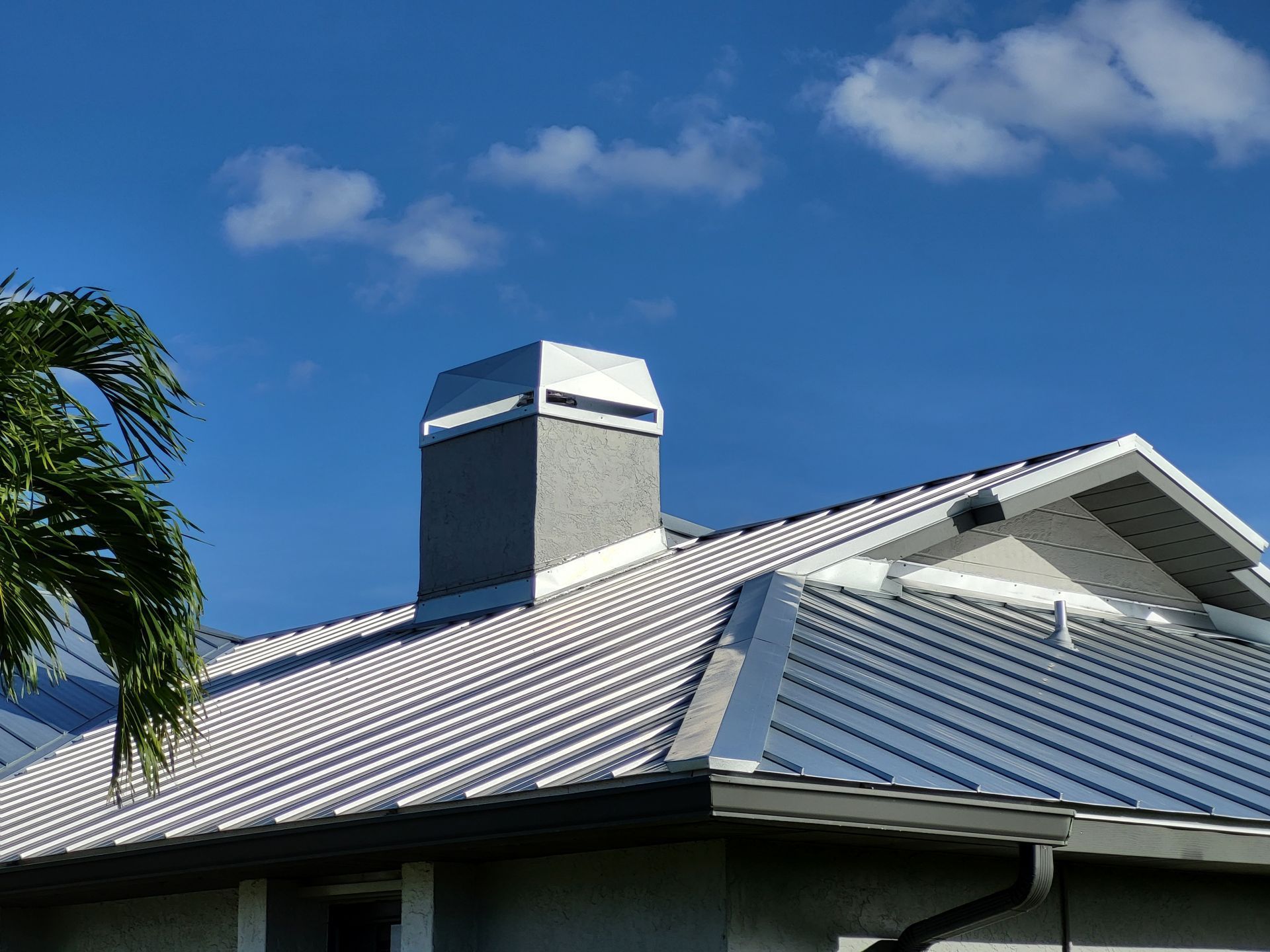 A metal roof with a chimney stack under a bright blue sky with a palm frond in the corner.
