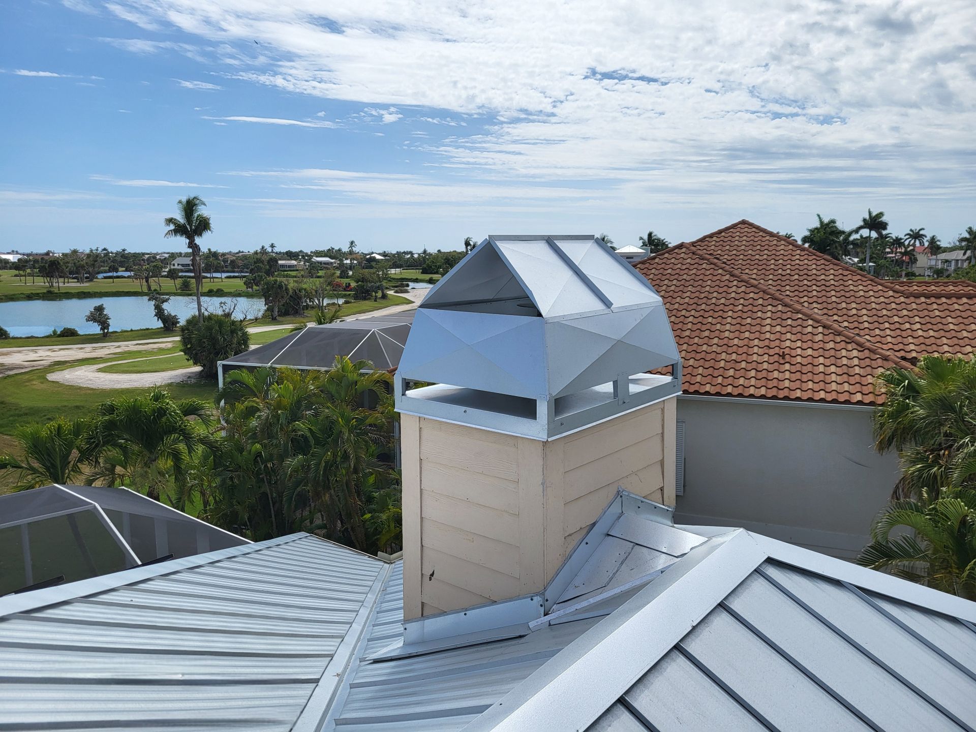 A metal chimney cap sits atop a light tan chimney on a metal roof, with a tropical landscape visible in the background.