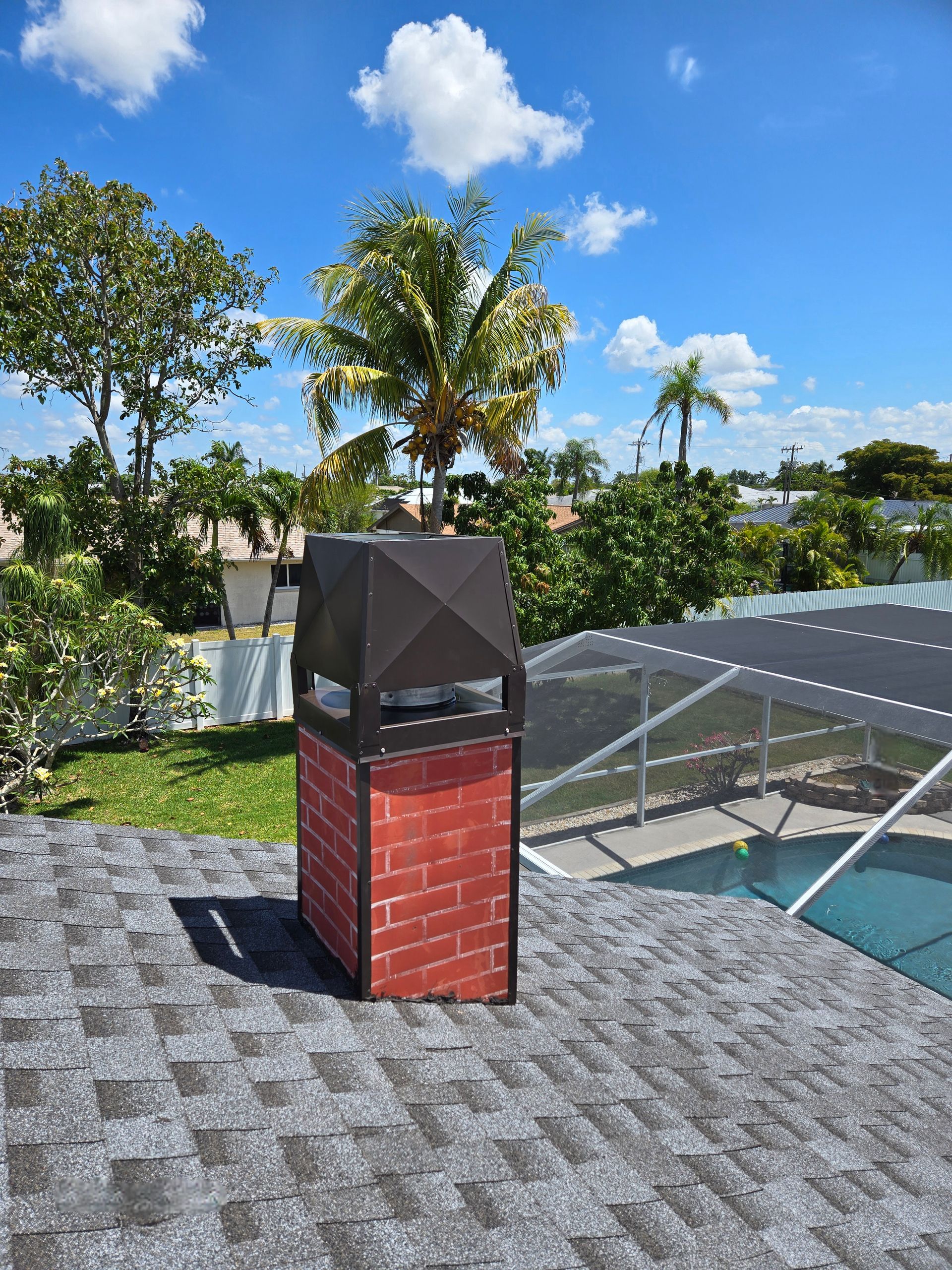 A brick chimney with a black metal cap on a gray shingled roof, set against a sunny, blue sky with palm trees.