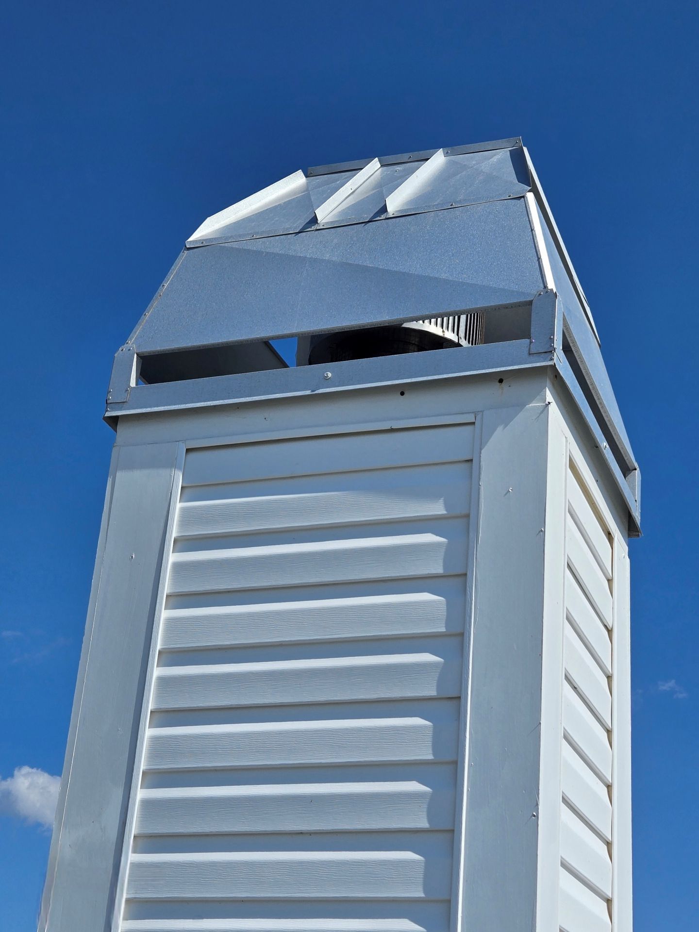 A white, louvered chimney stack topped with a geometric, metallic vent against a clear blue sky.