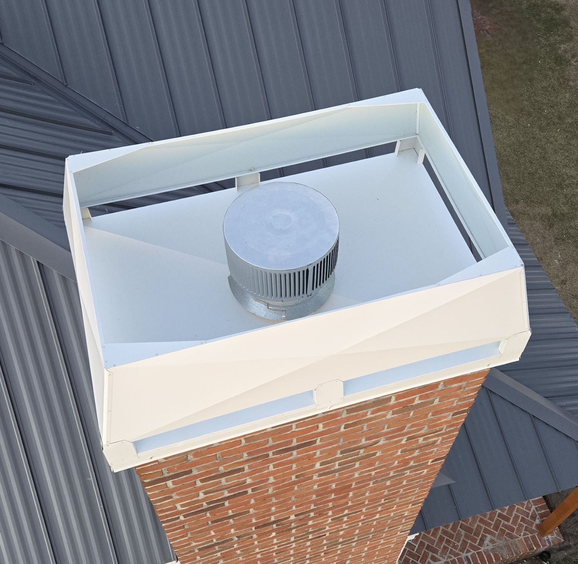 A white, rectangular metal chimney shroud with a central circular cap, mounted atop a brick chimney on a metal roof.