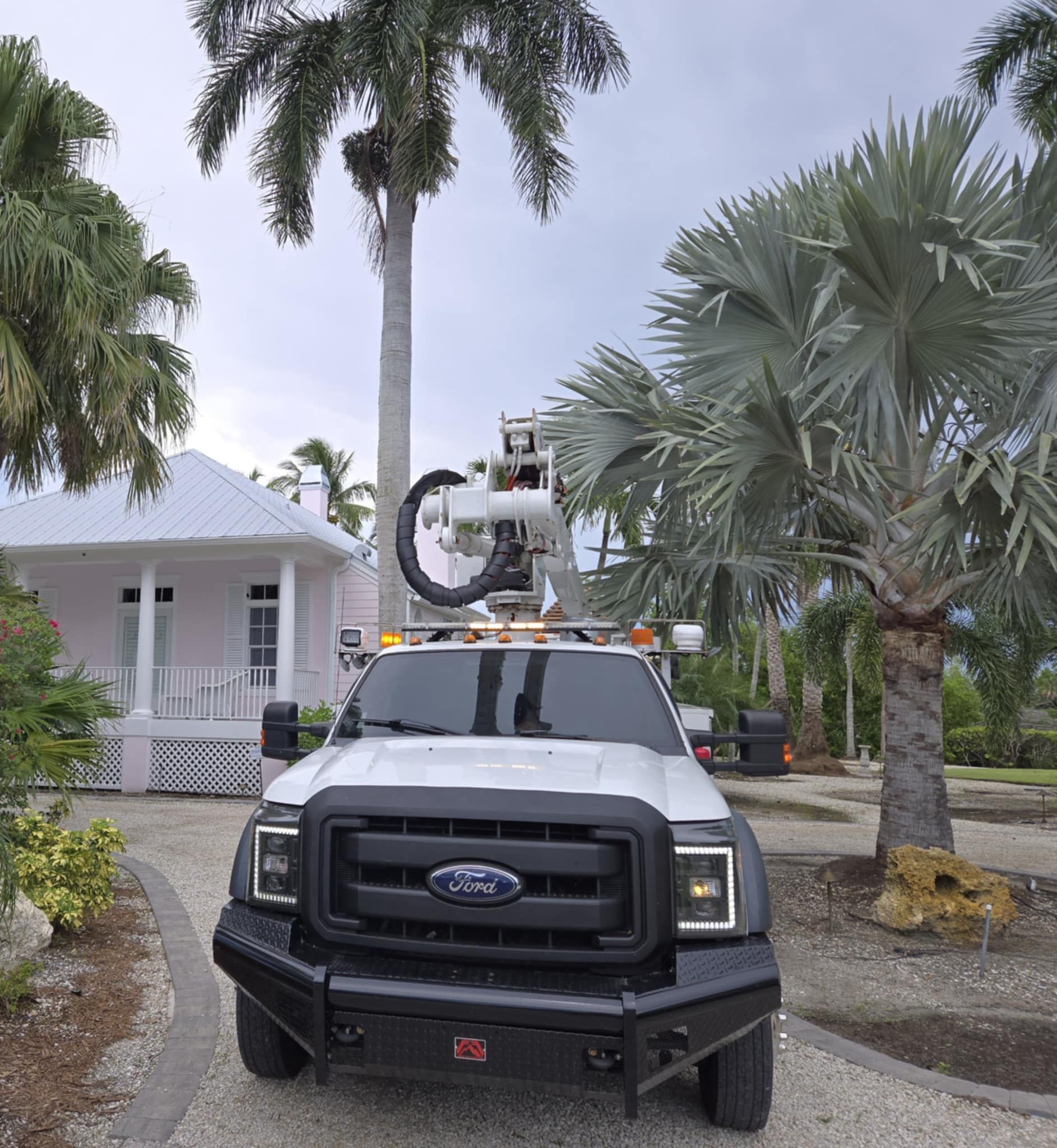 A white Ford utility truck equipped with a crane and camera rig parked on a gravel path near a house and palm trees.