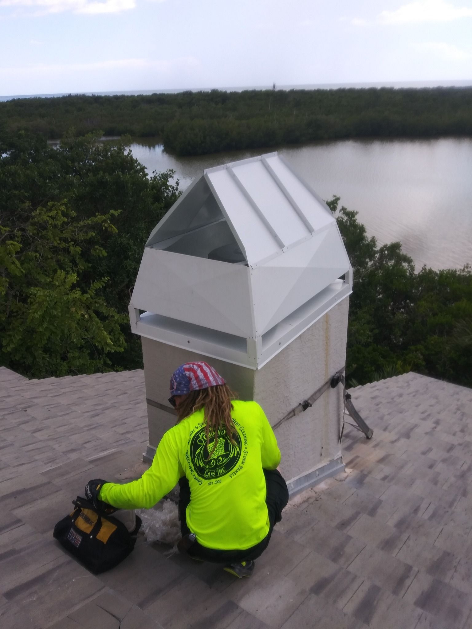A person in a neon yellow shirt works on a chimney cap on a roof overlooking a body of water and trees.