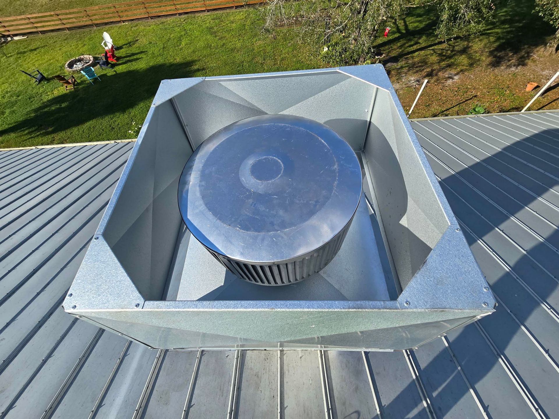 A metal chimney cap assembly installed on a gray standing-seam metal roof, viewed from above on a sunny day.