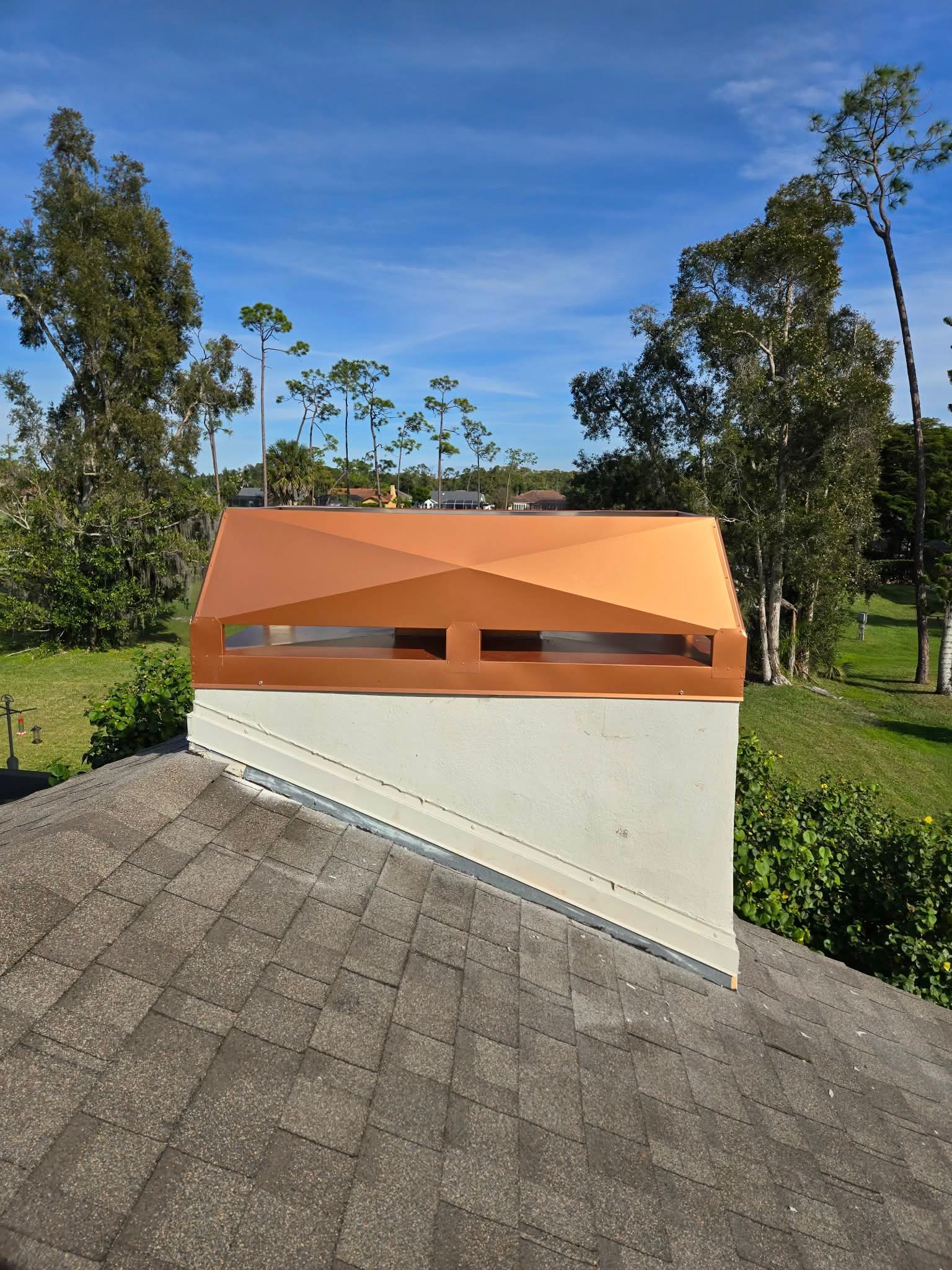 A copper-colored metal chimney cap sits atop a stucco chimney on a gray asphalt-shingled roof under a clear blue sky.