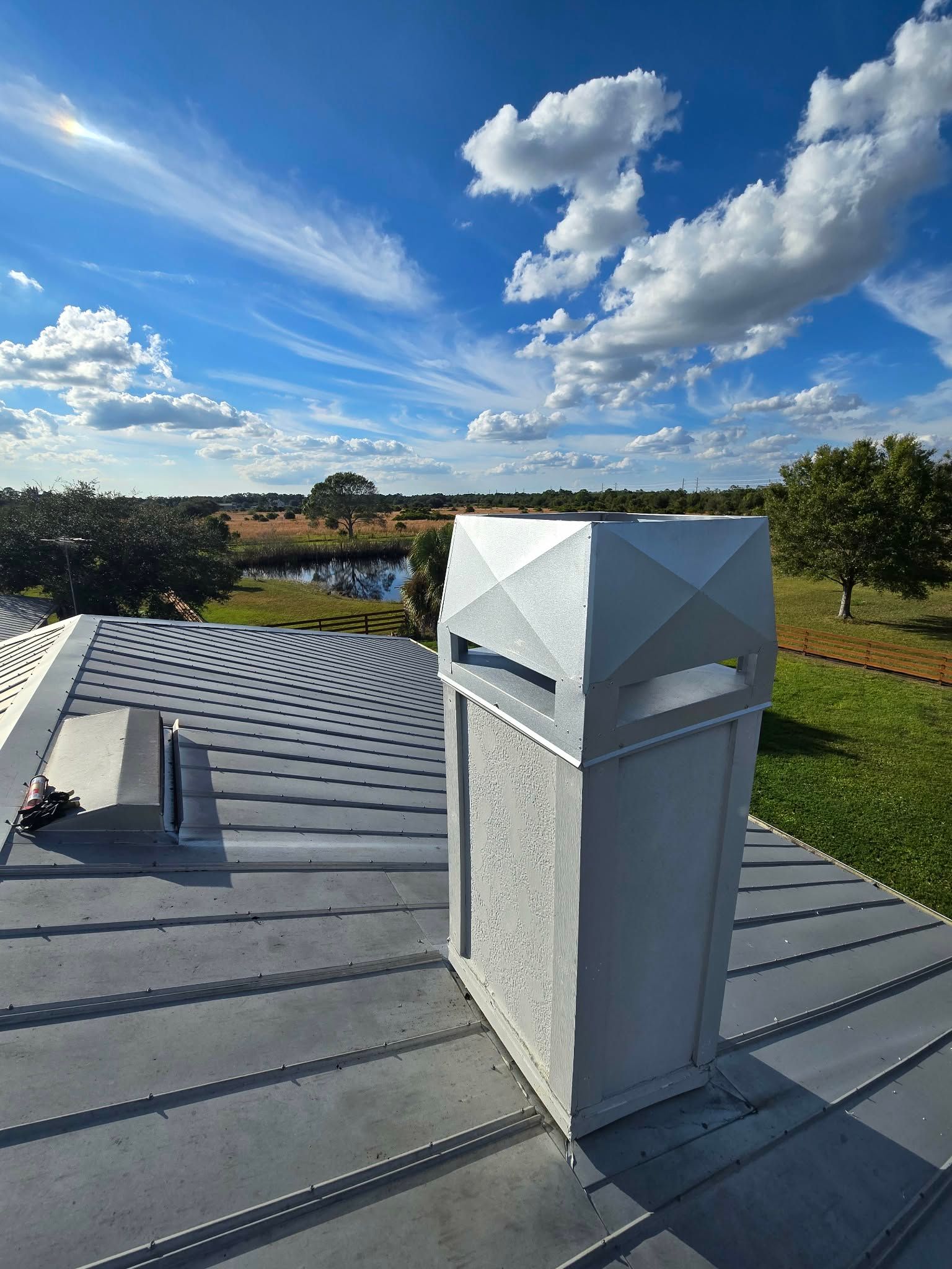 A white, rectangular chimney stack with a geometric cap sits on a grey metal roof under a bright, blue, cloudy sky.
