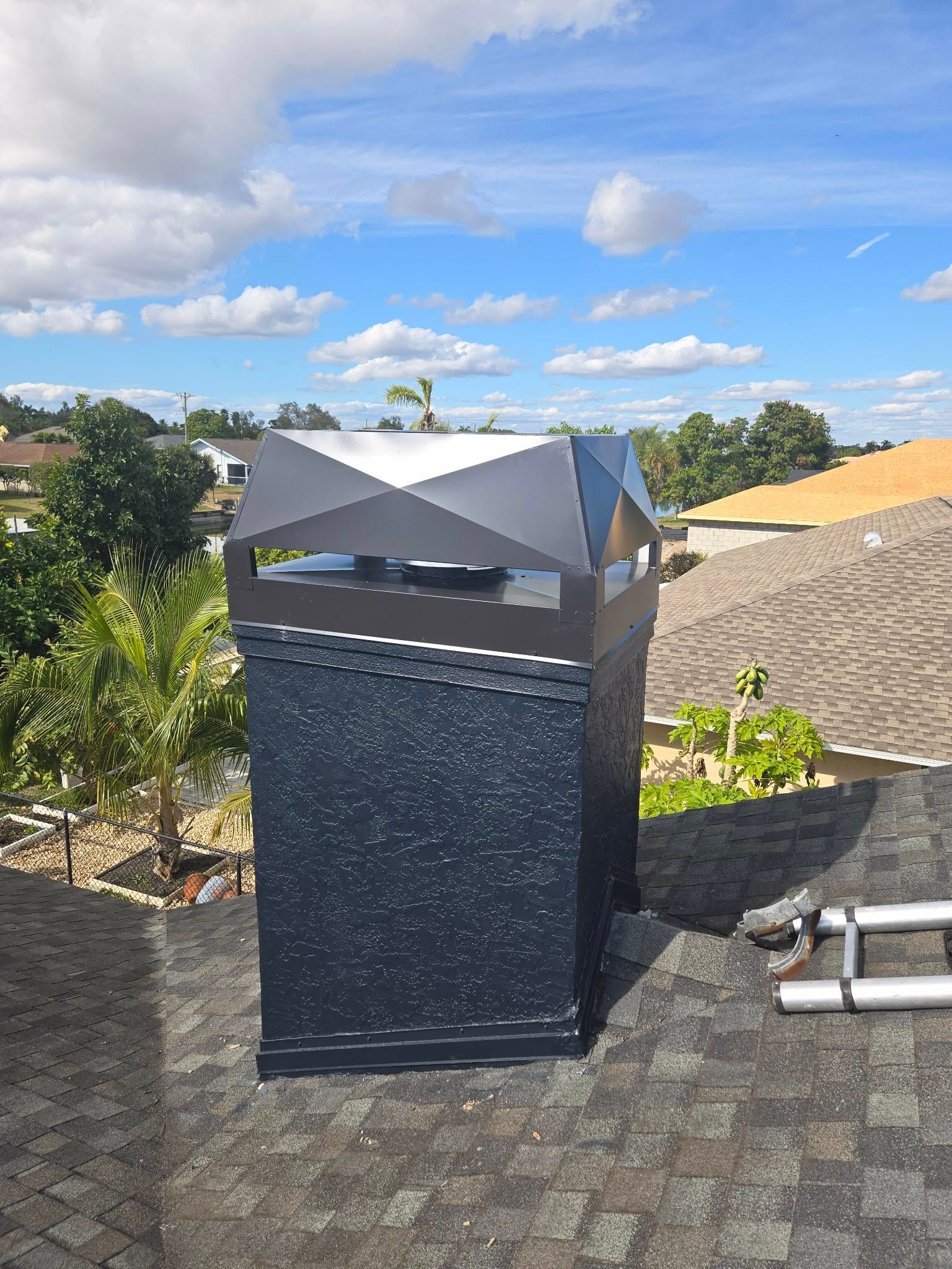 A dark gray chimney with a metal cap, situated on a shingled roof under a blue sky with scattered clouds.