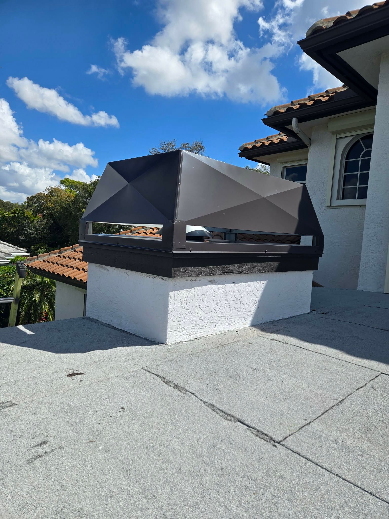 A dark metal chimney cap sits atop a white, textured masonry chimney base on a grey flat roof under a blue sky.