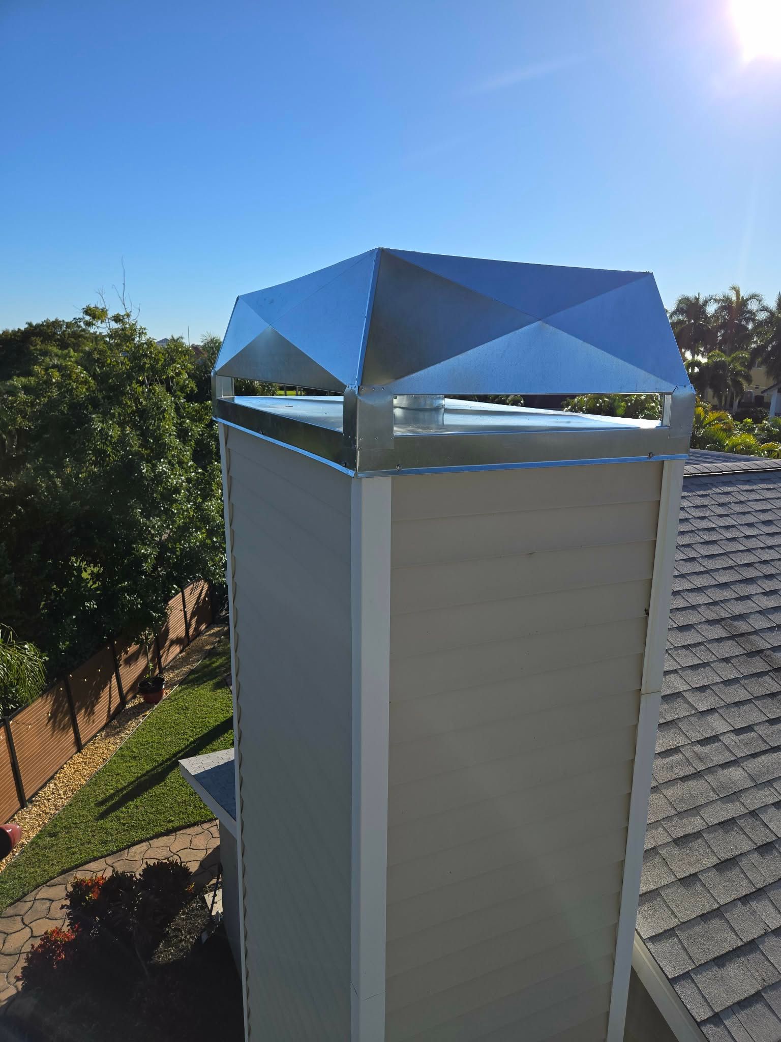 A tan-sided chimney with a shiny metal cap stands on a residential roof against a clear blue sky.