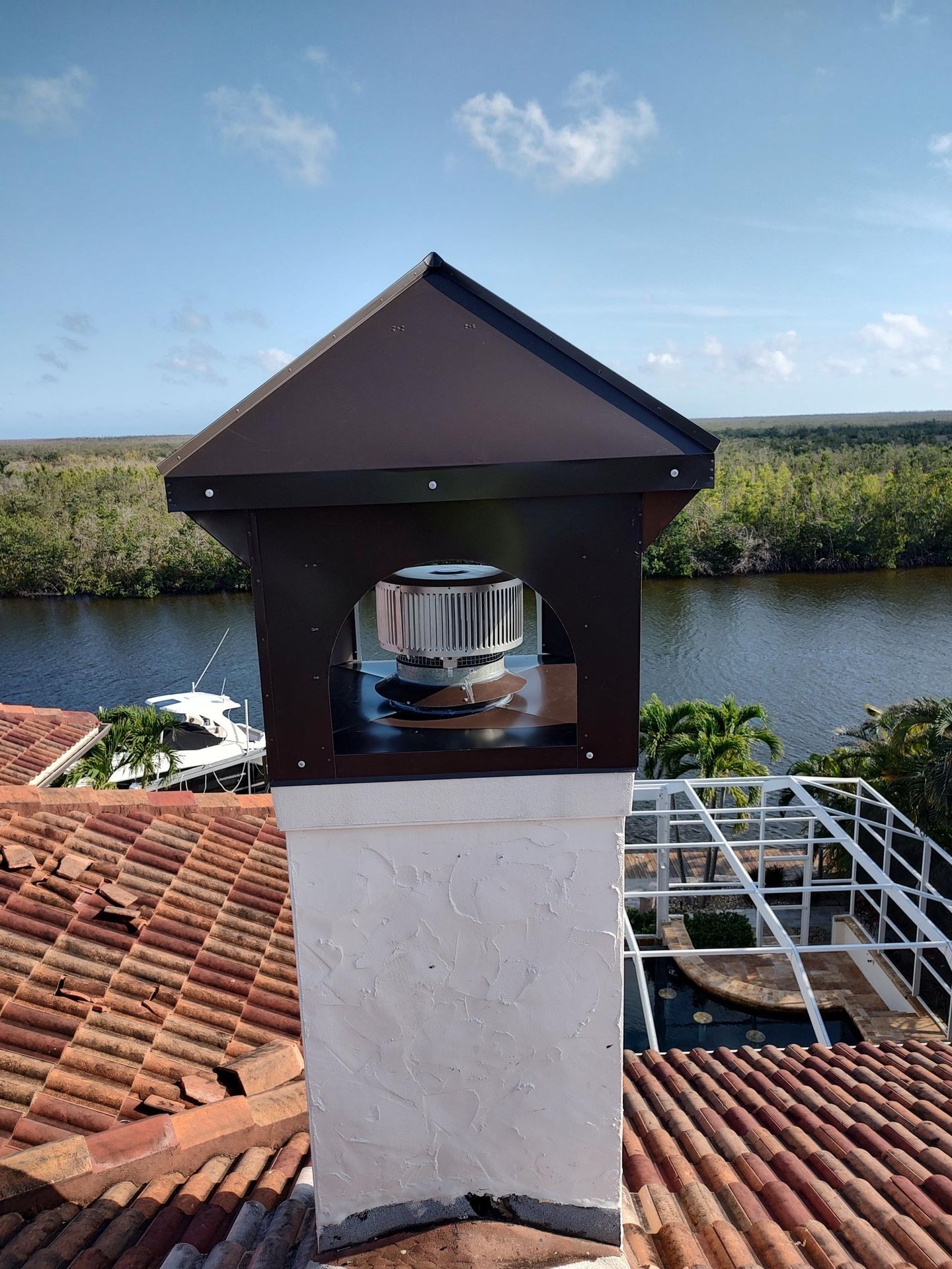 A metal chimney cap sits atop a white stucco chimney on a tiled roof overlooking a river and tropical landscape.