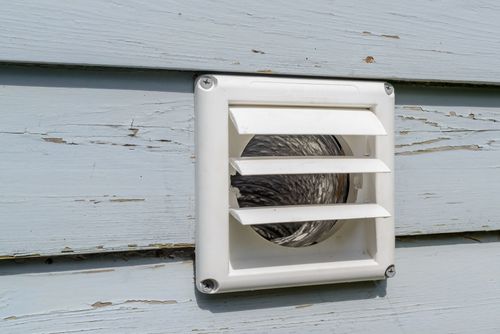 A white plastic dryer vent hood mounted on light blue horizontal wood siding.