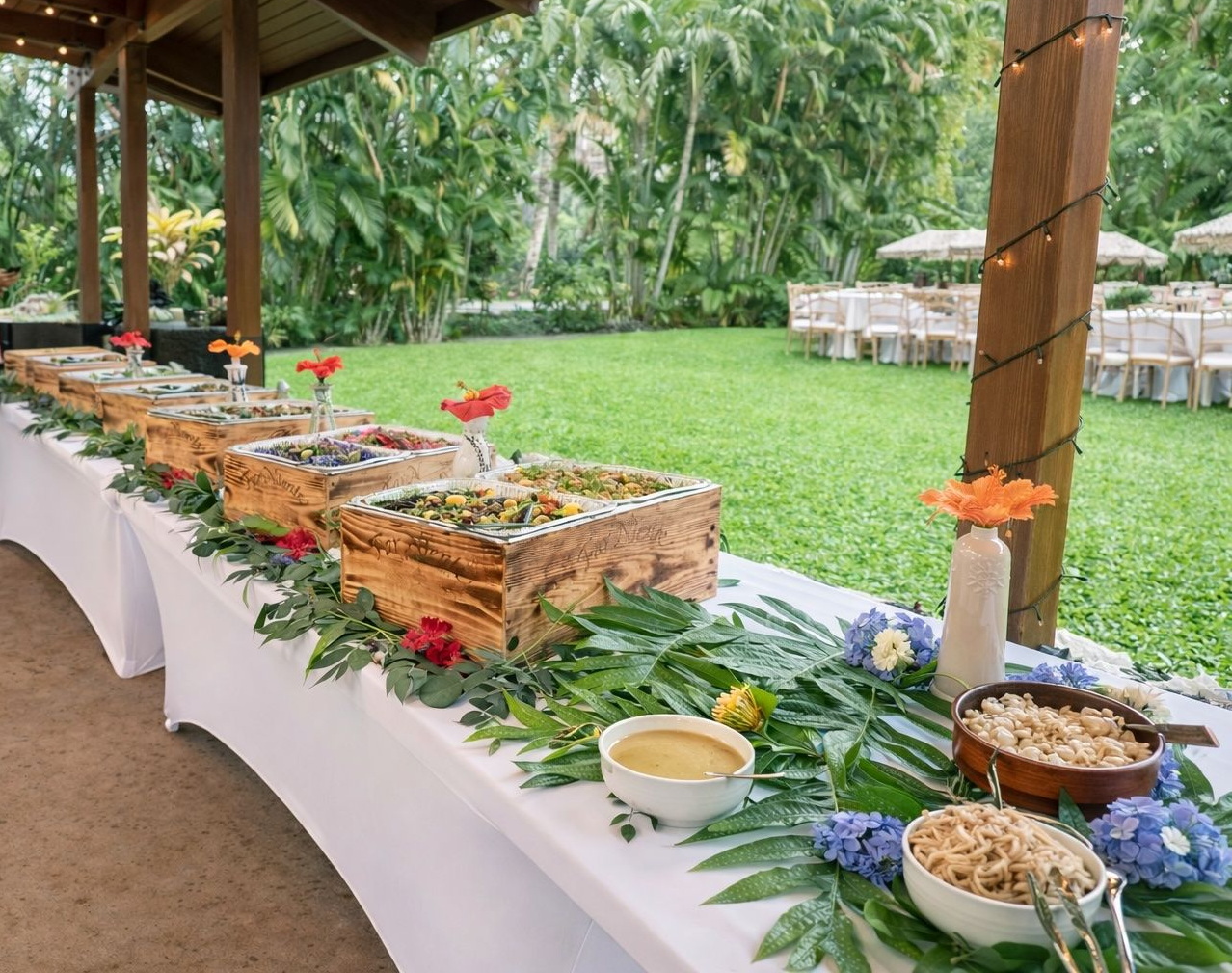 Outdoor buffet table with wicker baskets of food, white tablecloth, and tropical greenery in a garden pavilion