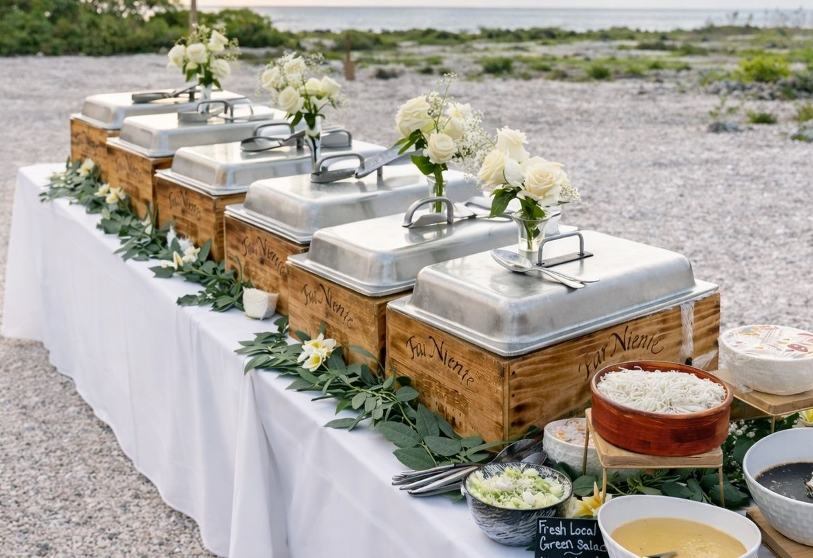 Buffet table with silver chafing dishes on a white cloth, decorated with greenery and white flowers outdoors