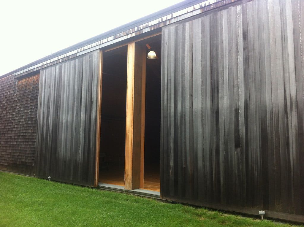 Black wooden building with an open doorway, seen from a grassy yard under overcast sky