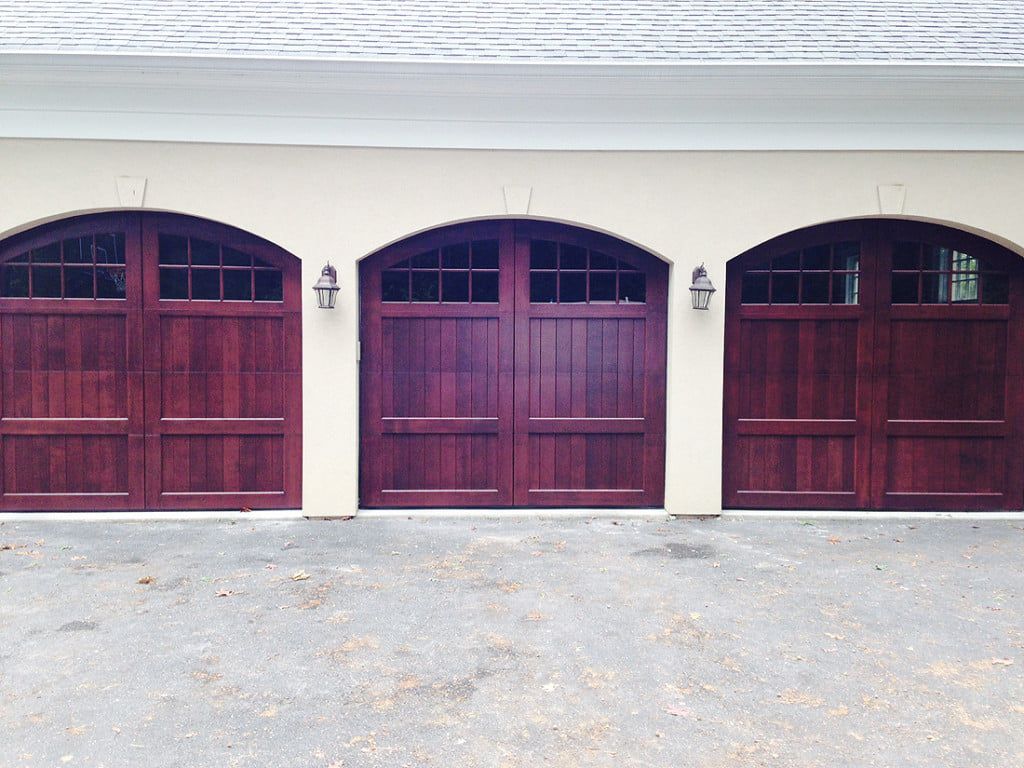 Three dark red garage doors on a beige house exterior with white trim and wall lanterns.