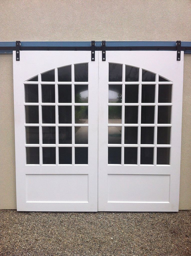 White double barn doors with arched window panes on a speckled floor against a beige wall.