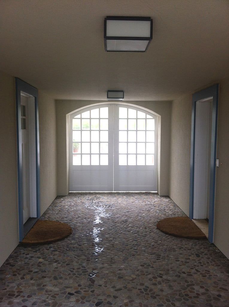 Empty hallway with double doors, tiled floor, and wall lights leading to a bright window.