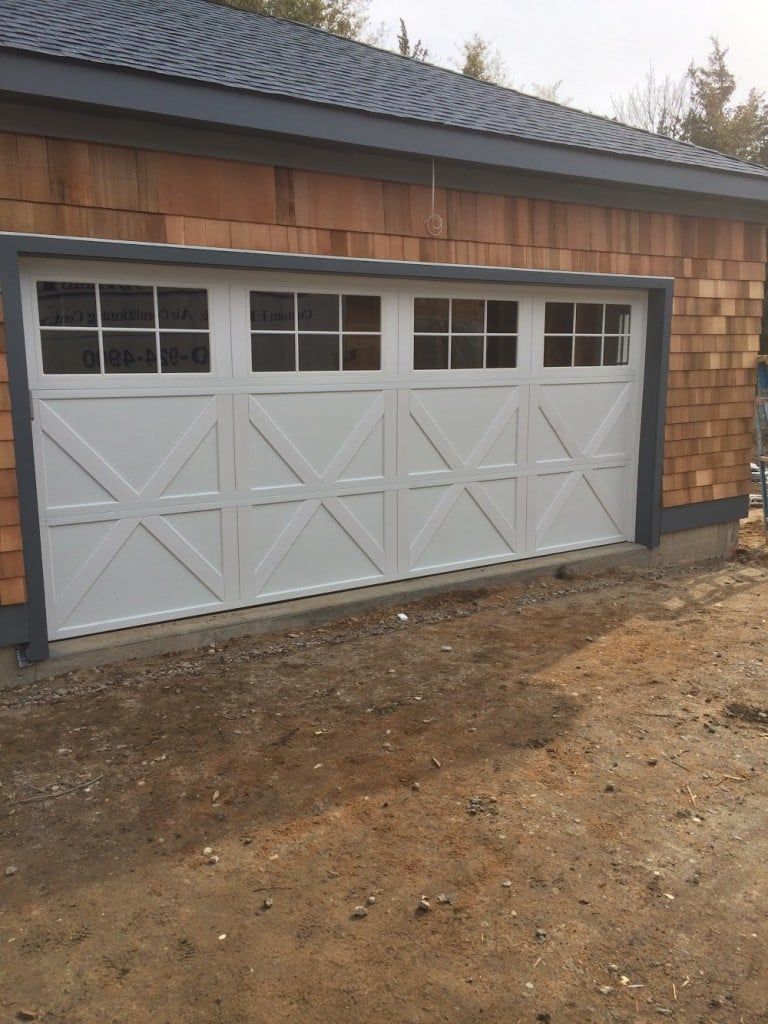 White garage door with small windows on a brown shingled building, viewed from a gravel driveway