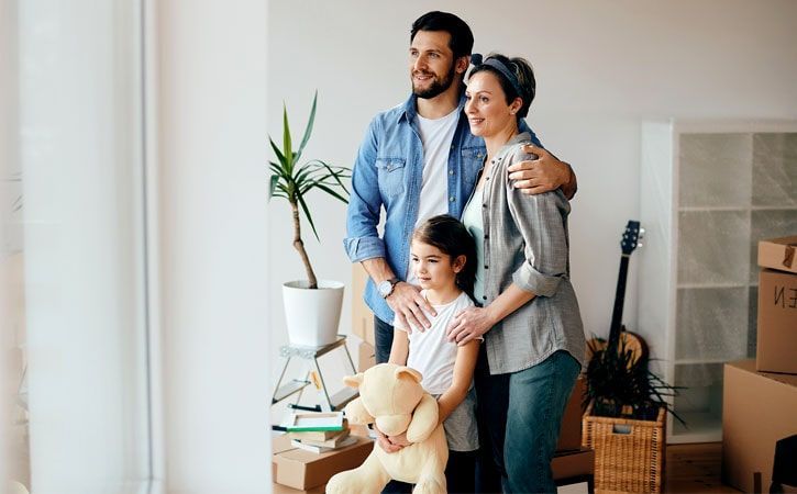 Familia de tres en su nueva casa, mirando por la ventana, sonriendo. Cajas y una guitarra de fondo.