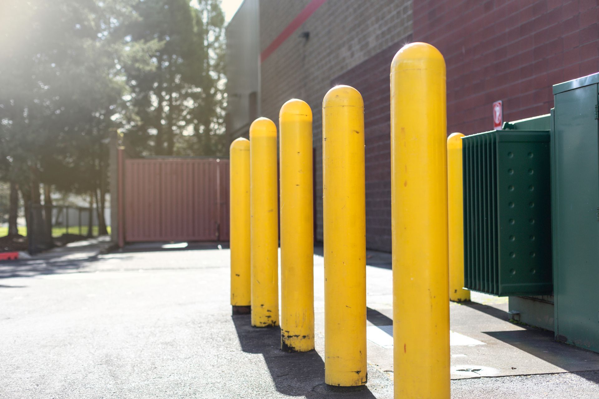 Yellow bollards protect a green electrical box, next to a brown metal container, in a paved area, with a building in the background.