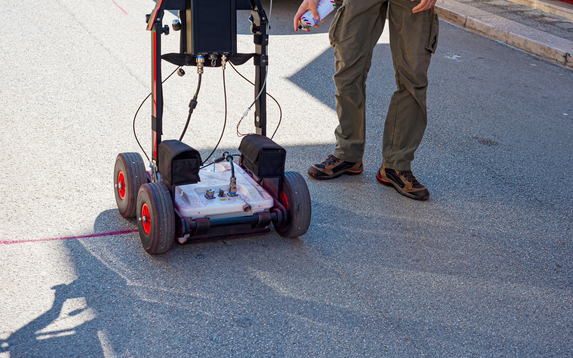 Person operating ground-penetrating radar on asphalt. The device is on wheels and has red accents.
