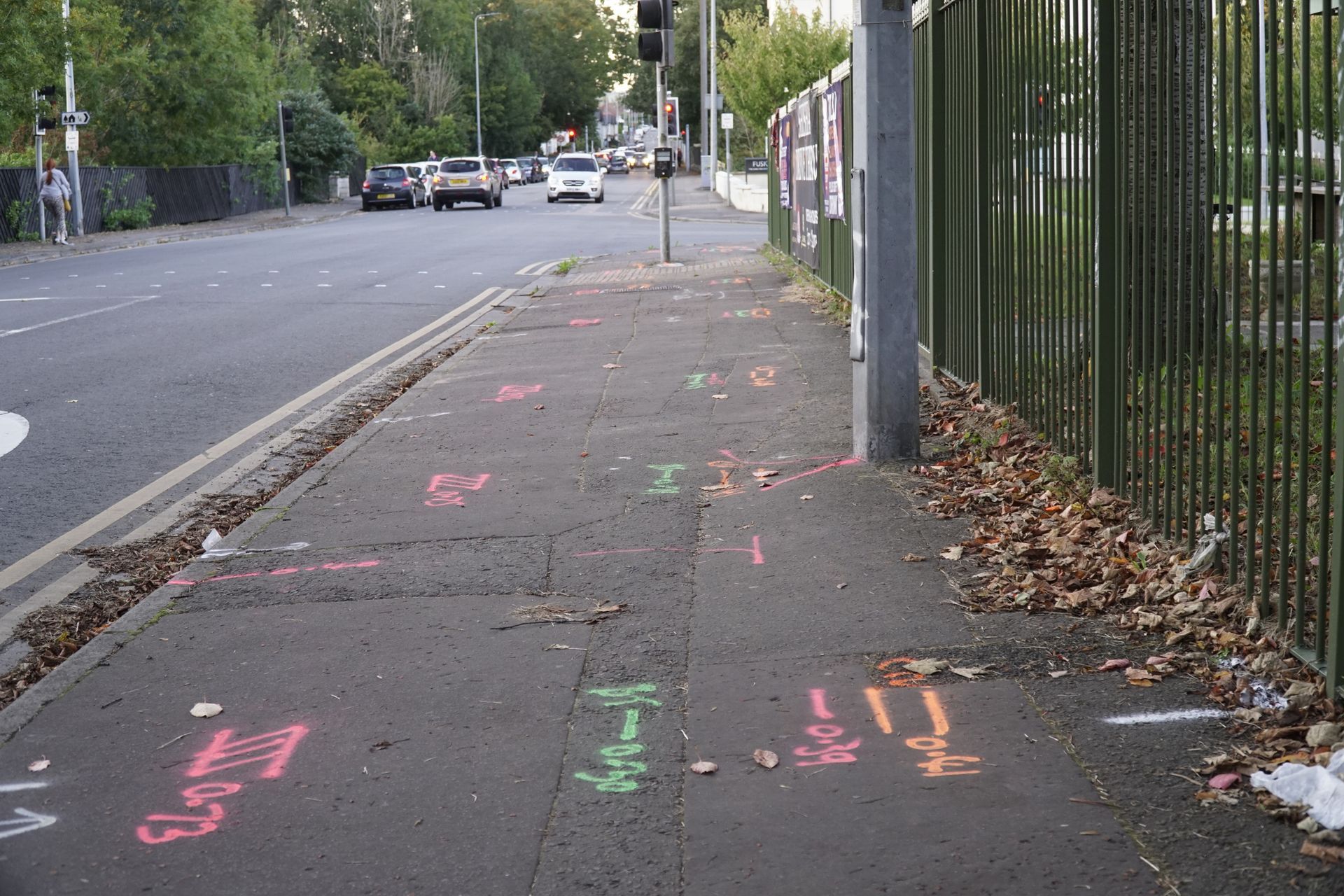 Sidewalk with utility markings. Cars travel on adjacent road. A metal fence borders the sidewalk.