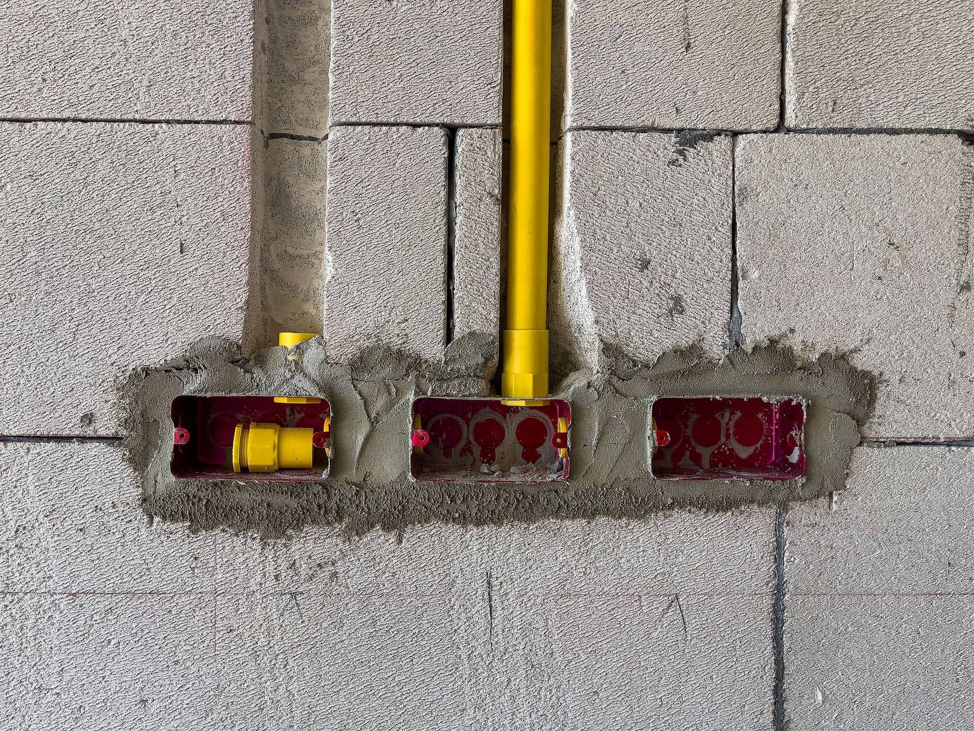 Electrical conduit and junction boxes embedded in a gray concrete block wall. Yellow conduit, red boxes.