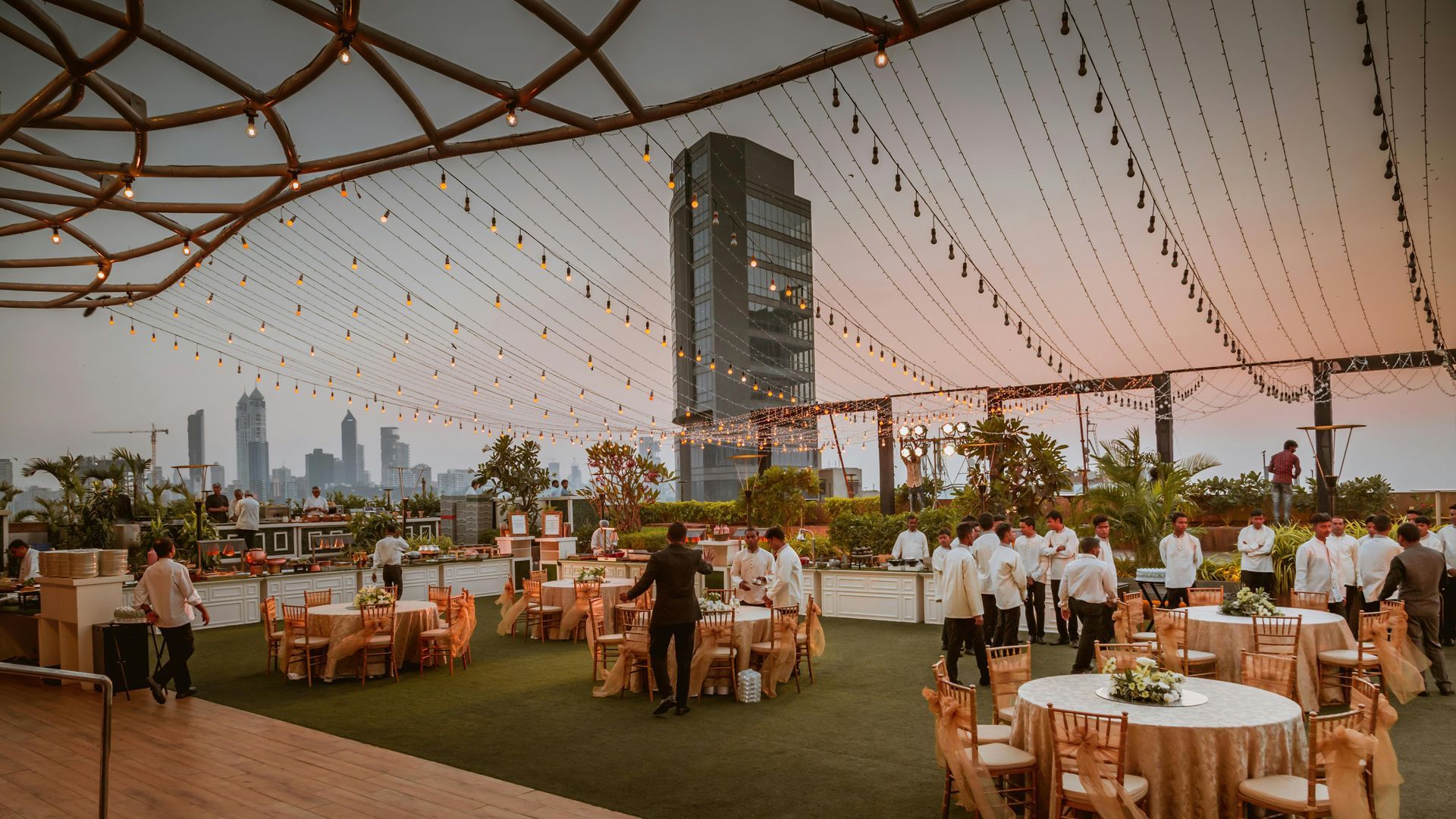Rooftop event with tables, servers, and city skyline at dusk, under string lights.