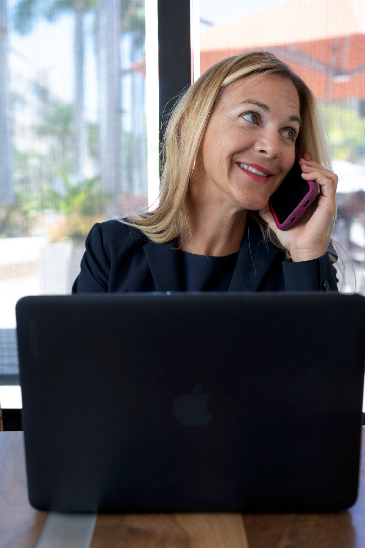Woman in a blazer smiles while talking on a cell phone and working on a laptop at a table near a window.