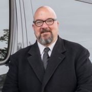 Smiling bald man with glasses in a black coat standing beside a vehicle