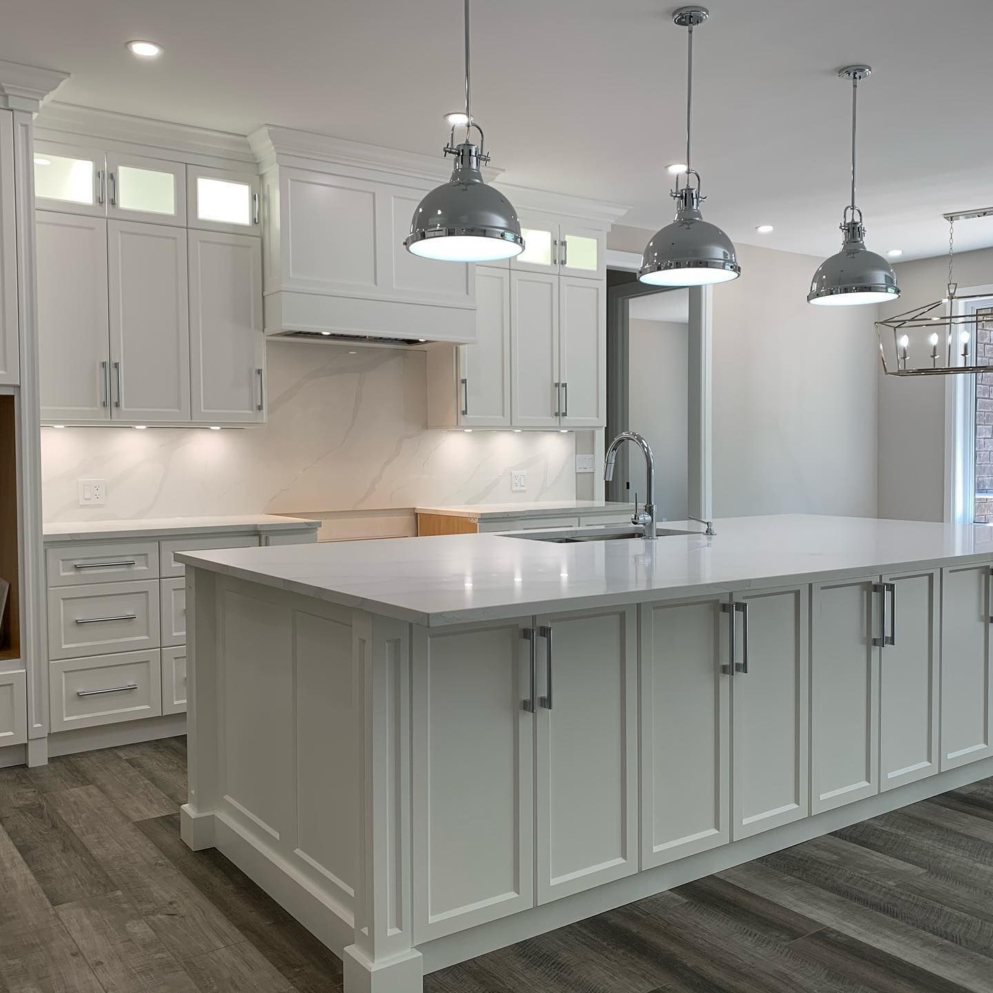 Modern white kitchen with a large island, marble-style countertops, three industrial pendant lights, and wood flooring.