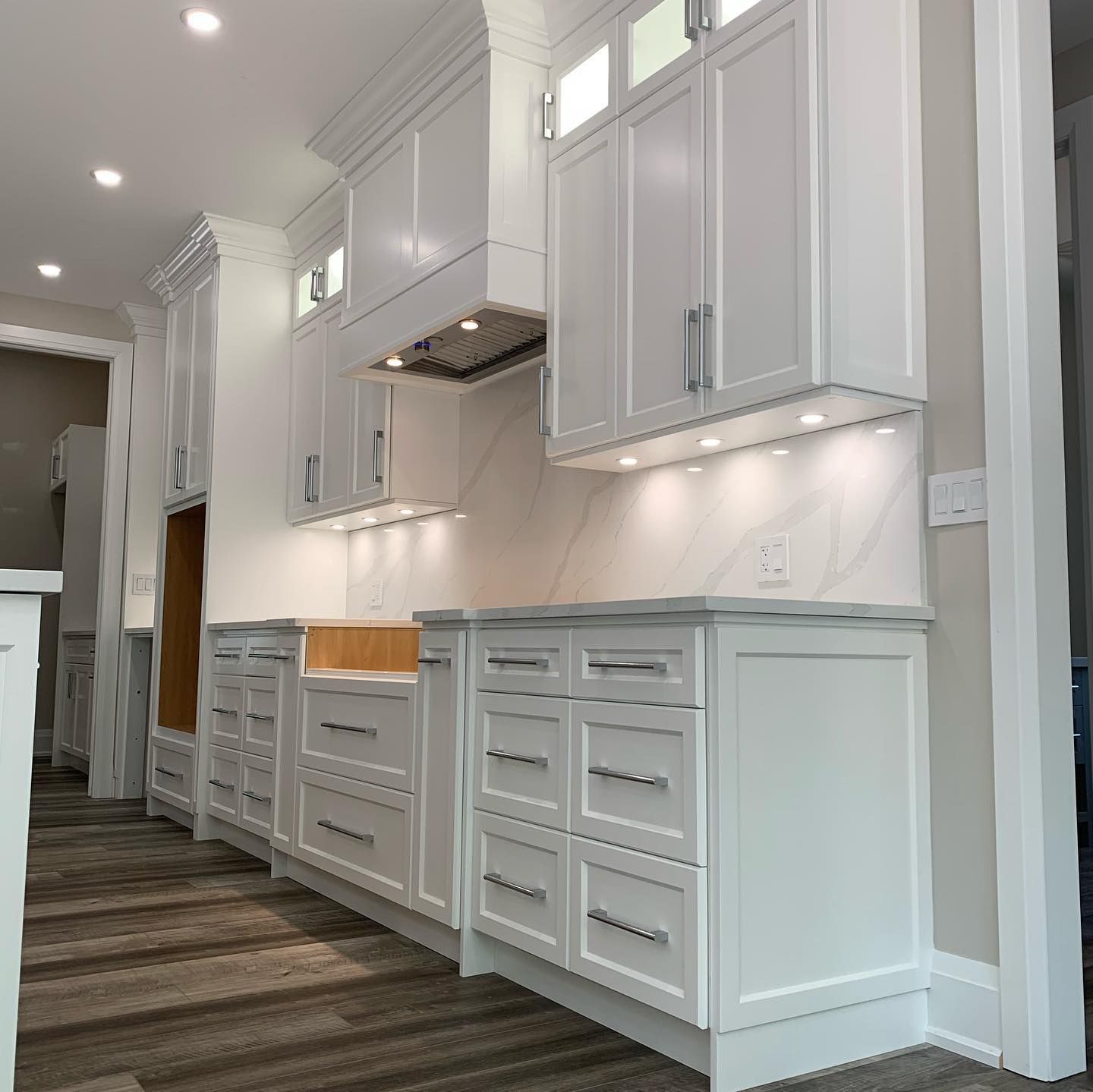 A modern kitchen featuring white cabinets, a farmhouse sink, under-cabinet lighting, and dark wood-look flooring.