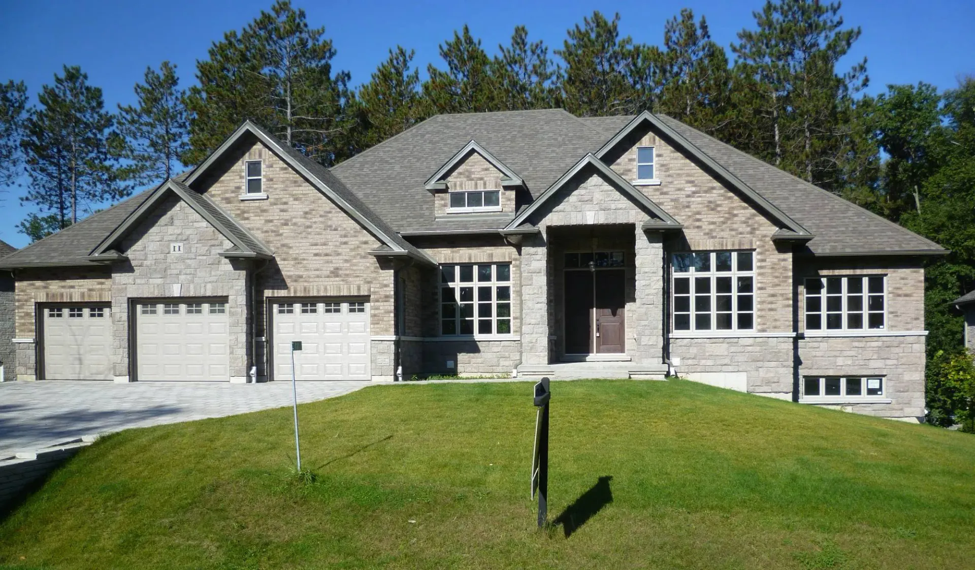 A large, single-story tan brick suburban house with a three-car garage and a grassy front lawn under a clear blue sky.