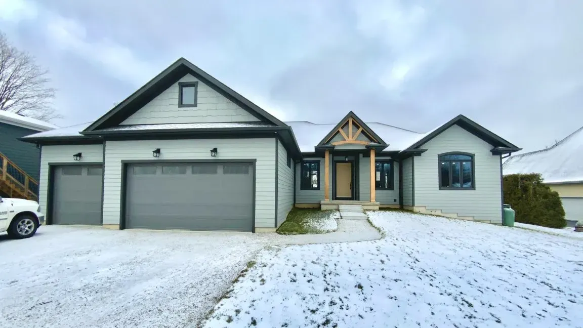 A light green single-story suburban house with a two-car garage under a snowy sky, seen from a snow-dusted front lawn.