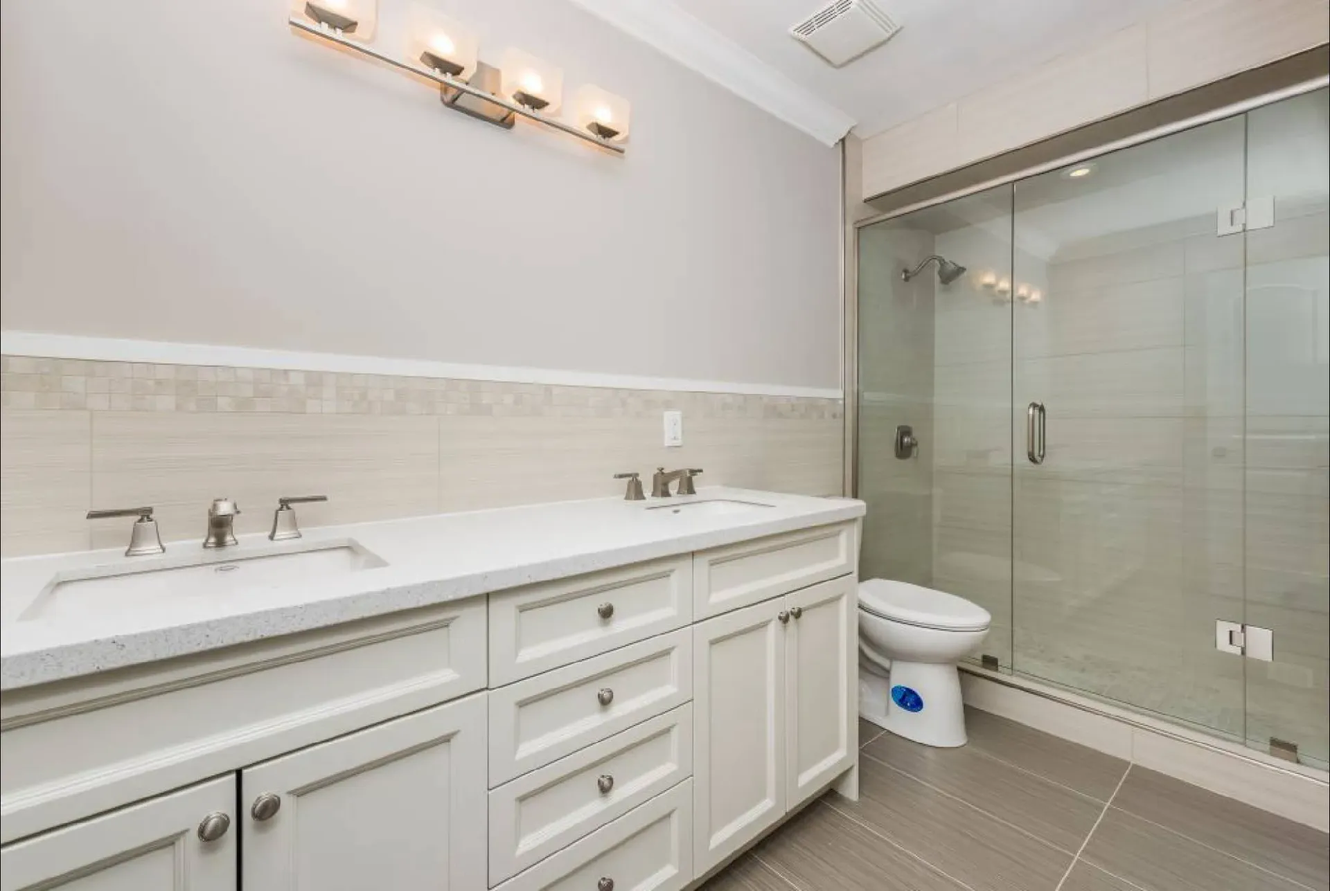 A modern bathroom featuring a white double vanity, light gray tile walls, and a glass-enclosed shower with a toilet.
