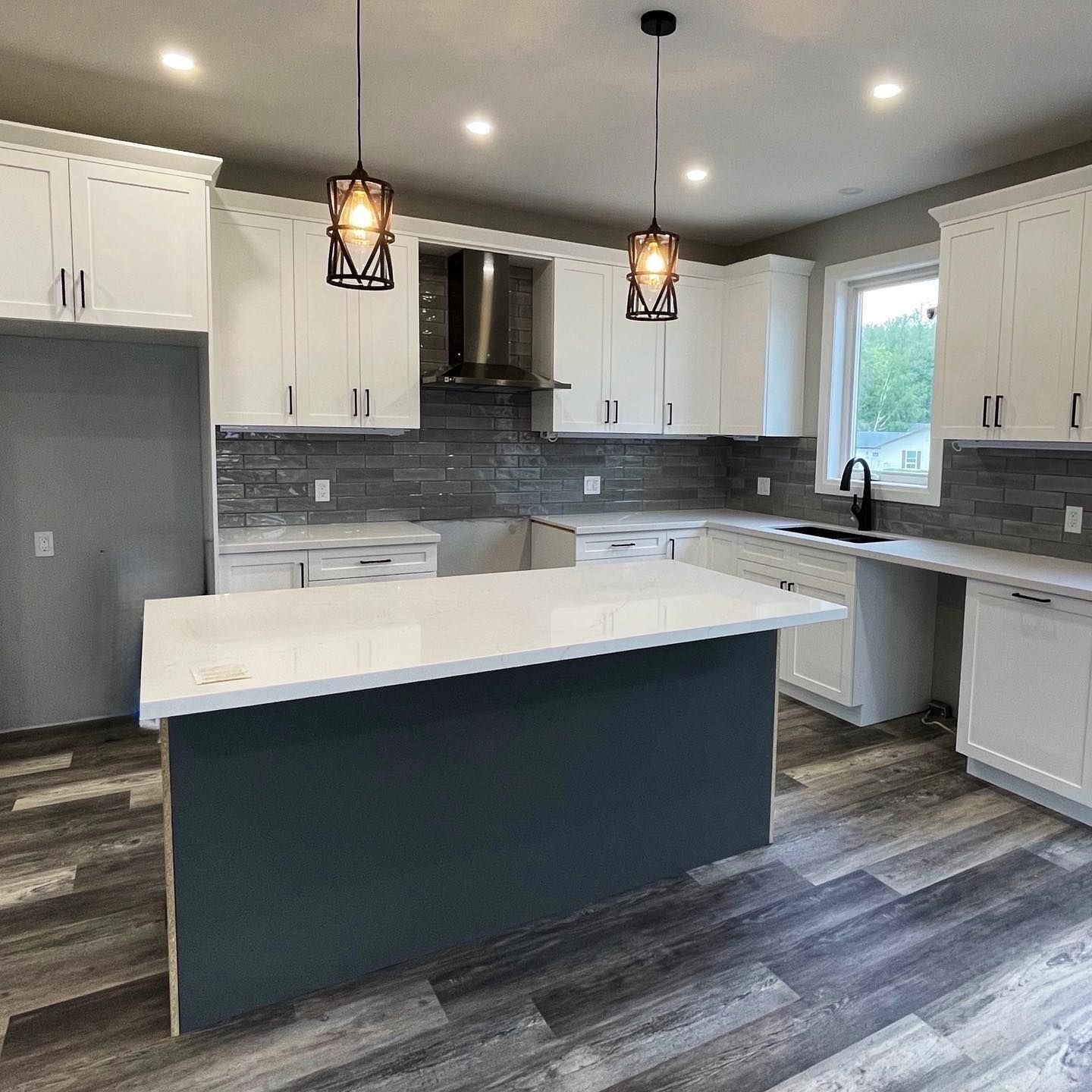 Modern kitchen with a dark blue island, white cabinets, grey stone backsplash, and two hanging pendant lights.