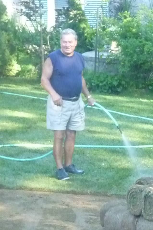 A person stands in a grassy yard, spraying a hose toward a pile of rolled sod while looking toward the camera.