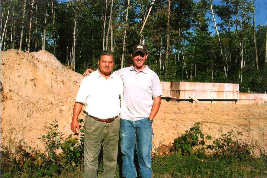 Two men standing in front of a dirt pile and building foundation at a construction site.