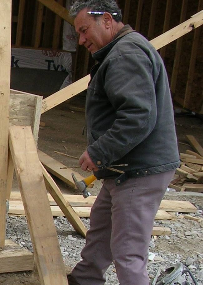 A construction worker in a grey jacket holds a hammer while working on a wooden frame at a building site.