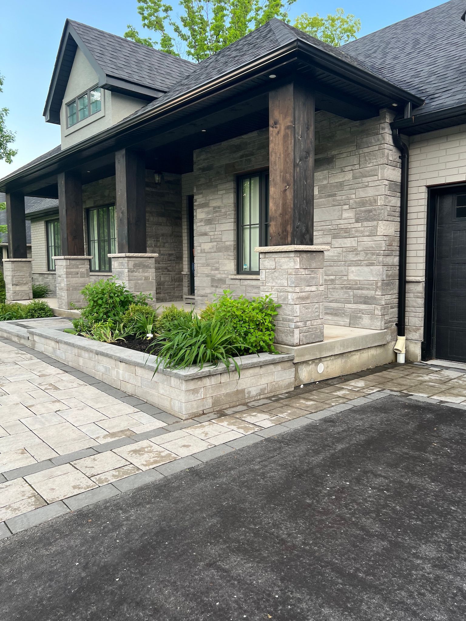 Stone-and-brick house entrance with dark roof, paved driveway, and small landscaped shrubs